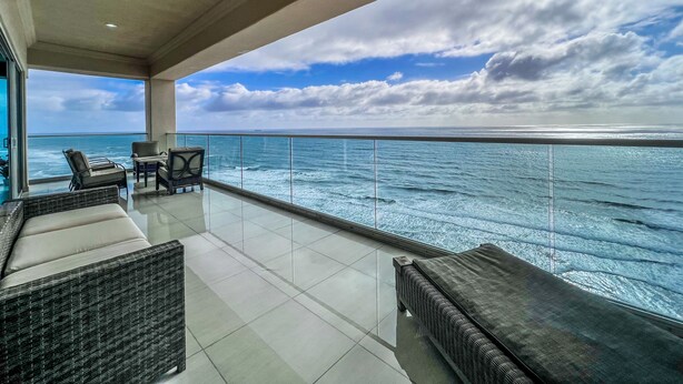 Un balcon avec vue sur l’océan, aménagé avec des canapés et des fauteuils en osier, et doté d’une rambarde en verre.