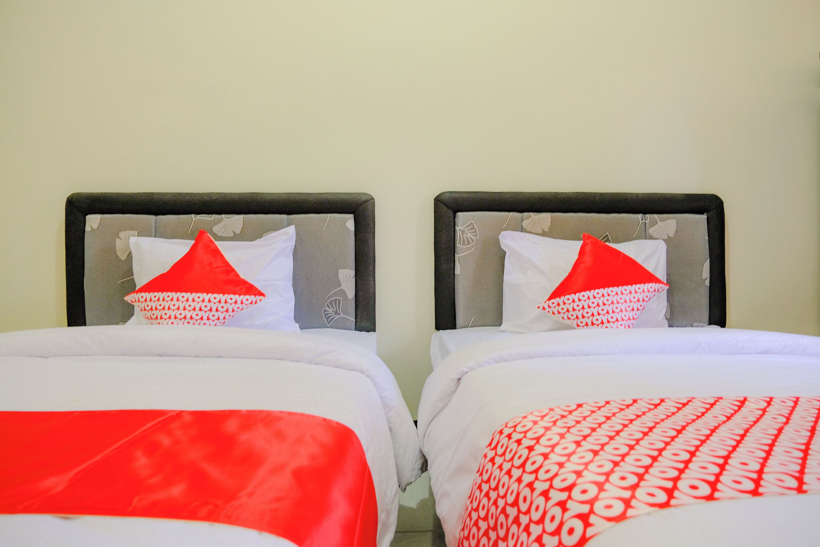 Dos camas con ropa de cama blanca y detalles rojos, ubicadas en una habitación con pared lisa y sin ventanas visibles.