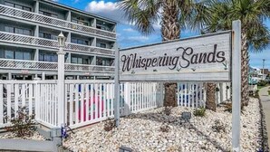 Een bord voor Whispering Sands, met een meerlaags gebouw op de achtergrond.
