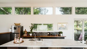 Une cuisine moderne dotée d’un grand îlot central, d’appareils électroménagers en acier inoxydable et offrant une vue sur le jardin grâce à de grandes fenêtres.