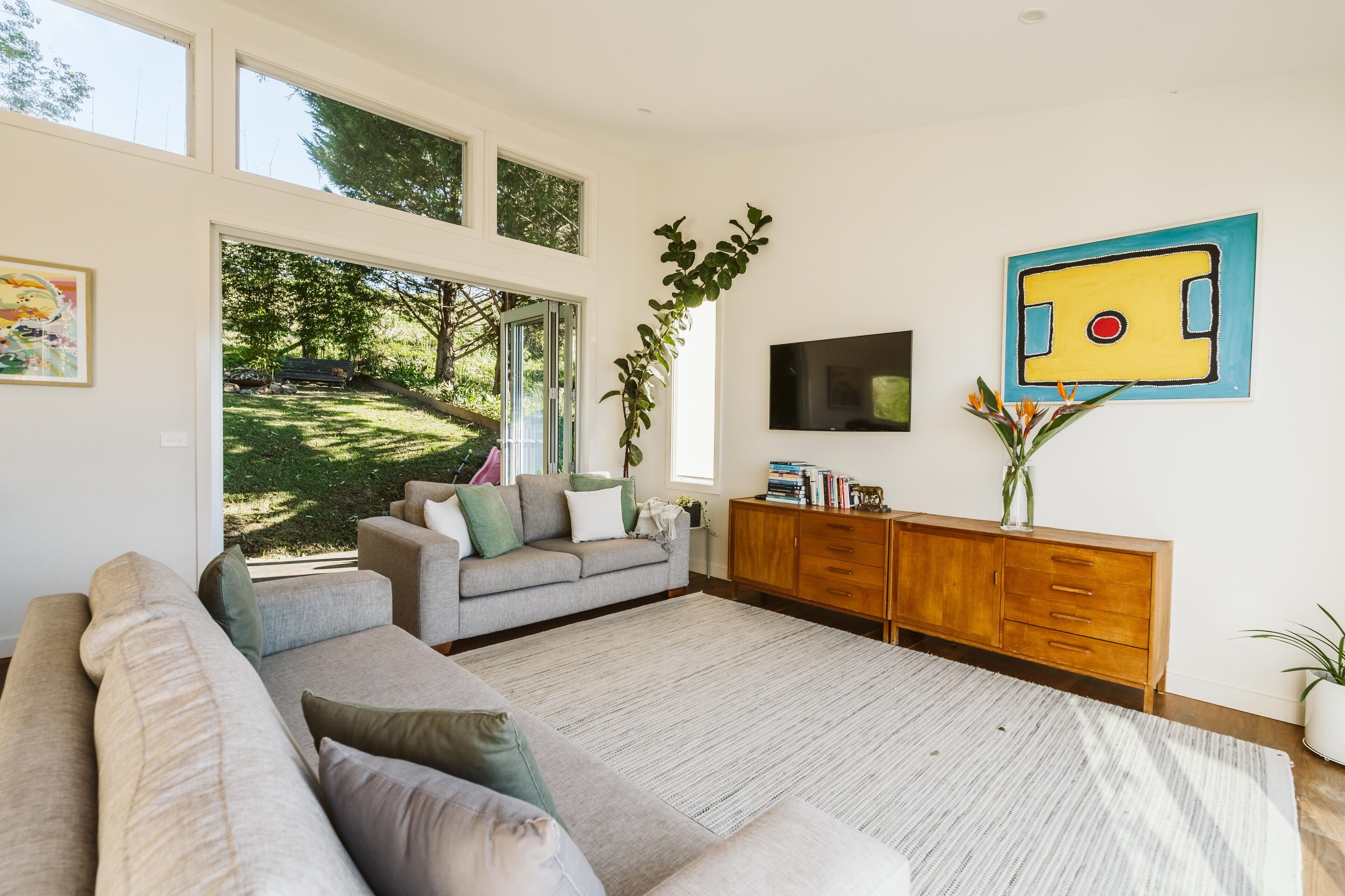 A modern living room with a large window, a grey sofa, a wooden cabinet, and a painting on the wall.