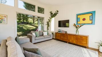 A modern living room with a large window, a grey sofa, a wooden cabinet, and a painting on the wall.