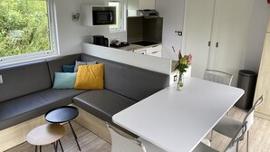 Una kitchenette compacta con microondas, cafetera y estufa. Cuenta con un comedor con mesa y sillas, y un sofá con cojines. Un ventanal proporciona luz natural.