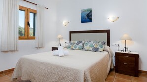 Una cama bien hecha con colcha, dos lámparas de noche, una mesita de noche, una ventana con cortinas y un cuadro colgado en la pared.