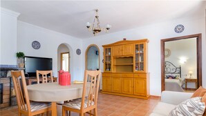 Una sala de estar con una mesa redonda, sillas de madera, un sofá, un televisor, un armario de madera y un piso de baldosas.