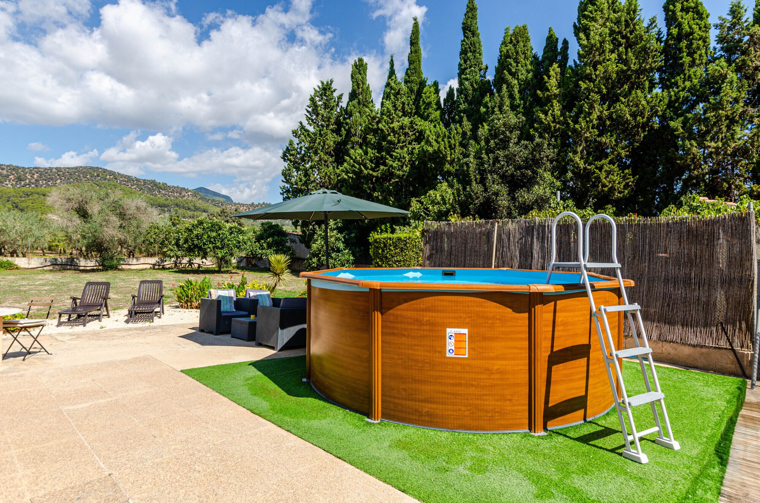 A wooden hot tub with a ladder, surrounded by a deck and chairs, with a wooden fence and trees in the background.
