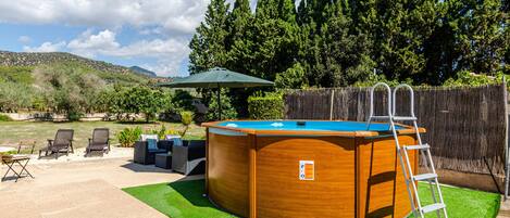A wooden hot tub with a ladder, surrounded by a deck and chairs, with a wooden fence and trees in the background.