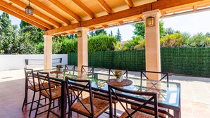 Un patio exterior con mesa y sillas de vidrio, vigas de madera y vistas a un seto cuidado y árboles.