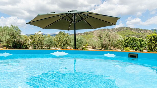 Una piscina con una sombrilla grande, rodeada de árboles y colinas al fondo.