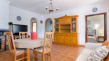 Una sala de estar con una mesa redonda, sillas de madera, un sofá, un televisor, un armario de madera y un piso de baldosas.