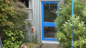 Een klein, afgesloten gebouwtje met een blauwe deur, omringd door weelderige begroeiing.