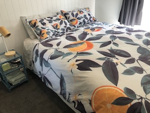A bed with a floral and fruit pattern bedspread, a bedside table with a lamp, and a small chair.