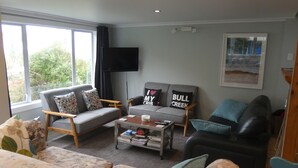 Een woonkamer met een grijze bank, een zwarte leren fauteuil en een gestreepe bank. Er staat een salontafel met boeken en een afstandsbediening op. Een flatscreen televisie is aan de muur bevestigd.