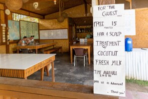 A wooden interior with a sign offering free hair and scalp treatment, coconut, and fresh milk. Tables and chairs are visible. - Where2next Hostel (El Nido)