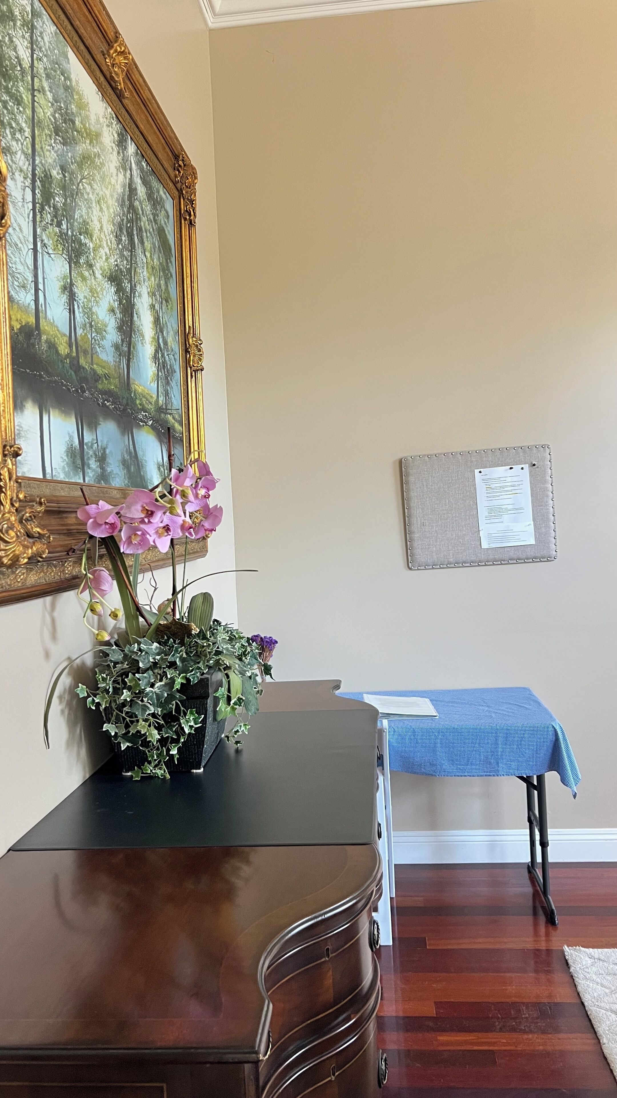 Une pièce comprenant une commode en bois, un vase avec des fleurs, un tableau encadré et un tableau d’affichage au mur.