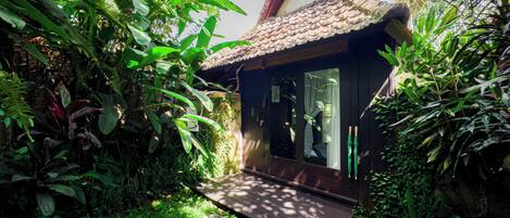 A small, rustic house with a tiled roof, surrounded by lush greenery and a wooden deck.