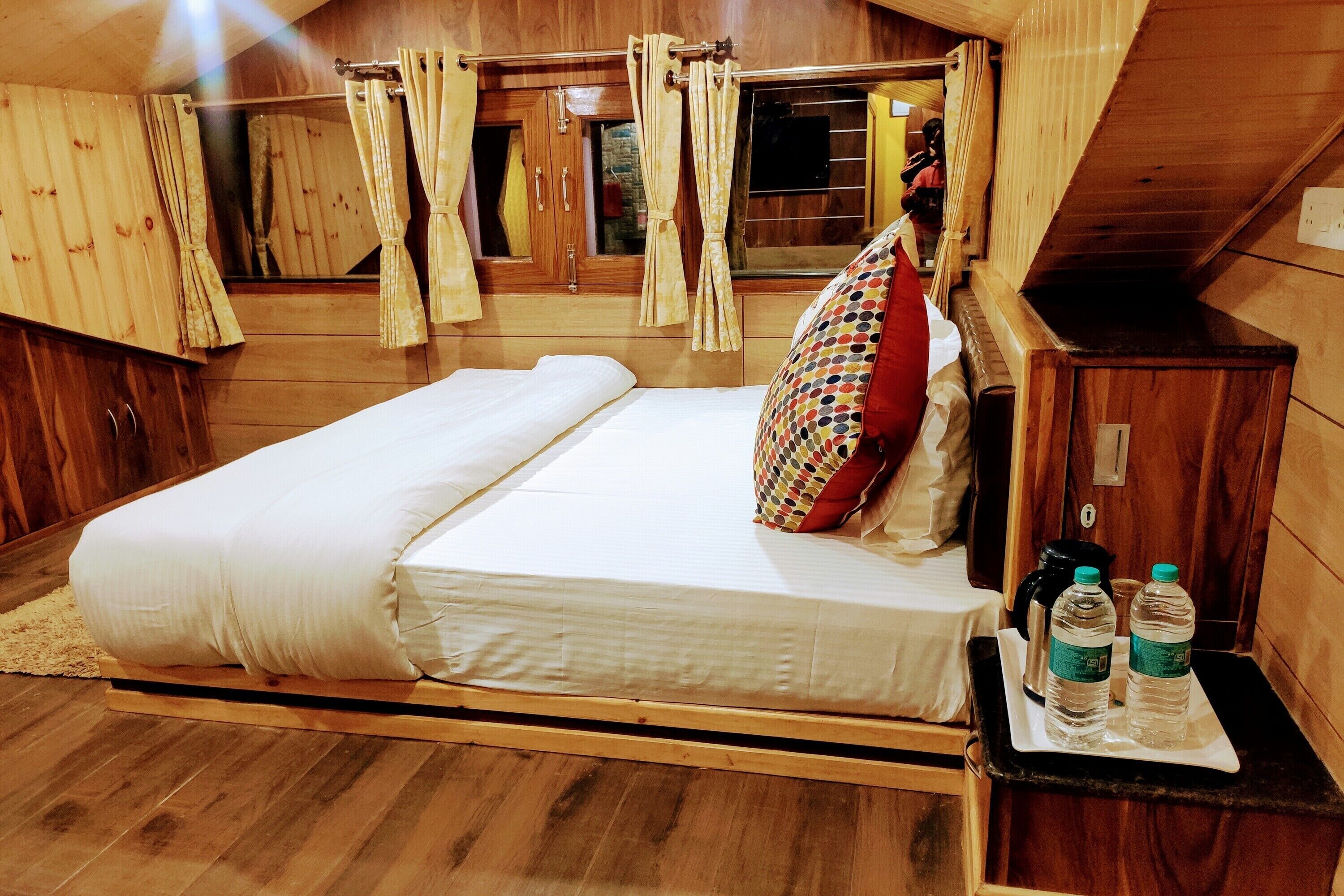 A wooden room with a bed, a nightstand with water bottles, and curtains.