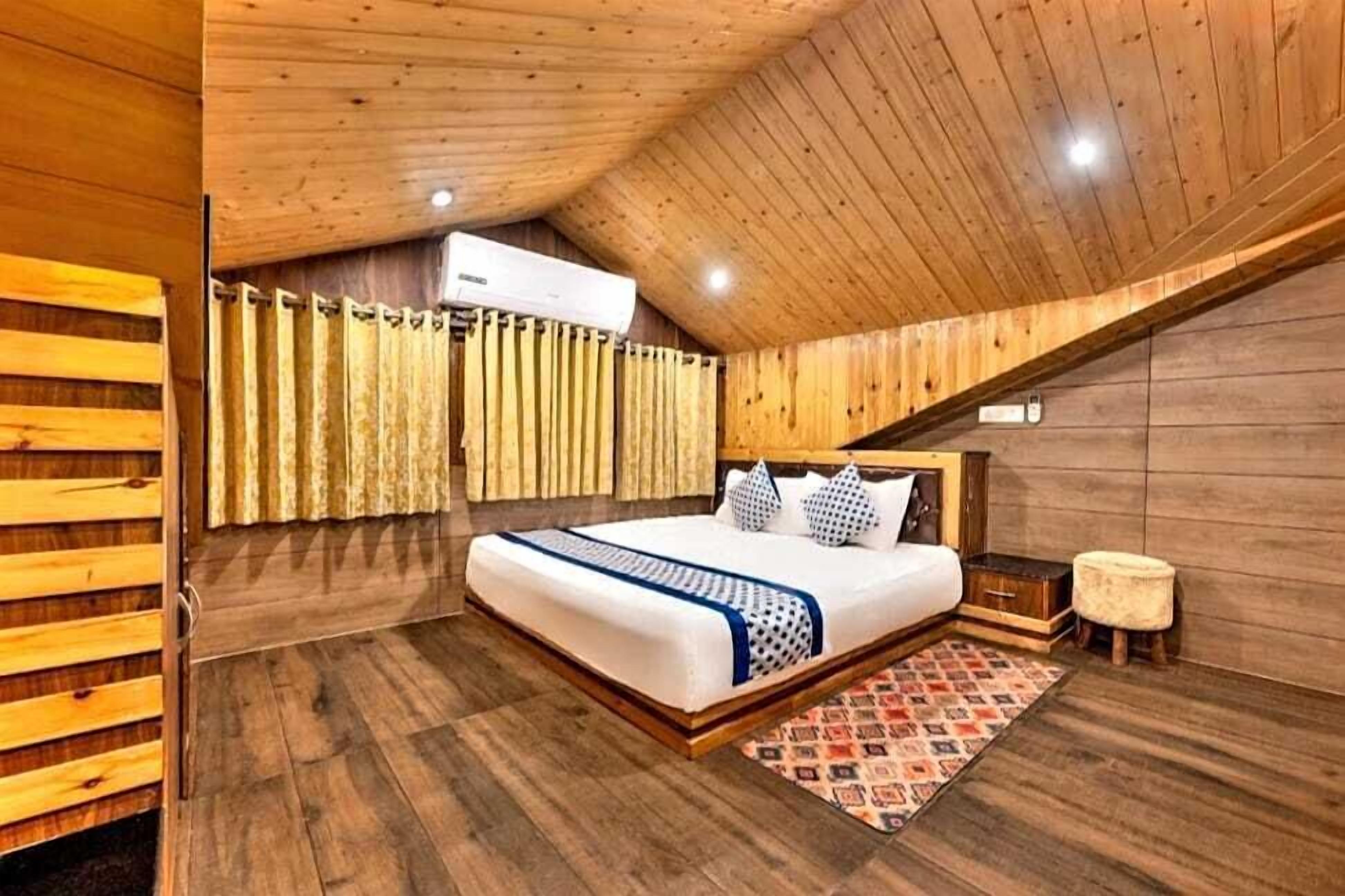 A bedroom with a wooden slatted ceiling, a bed with a patterned bedspread, a small stool, and a patterned rug.