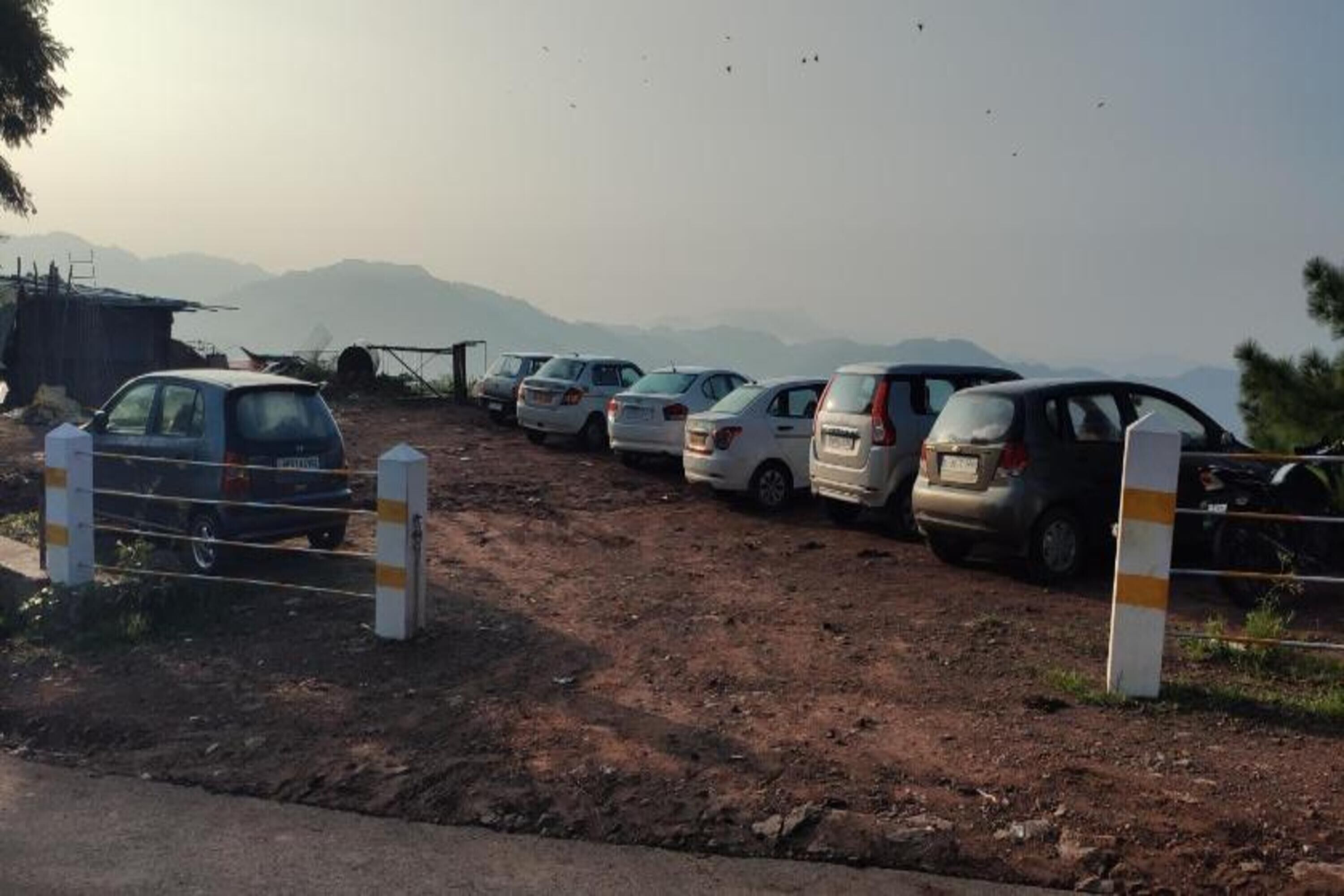 A parking area with several cars parked in a row, surrounded by a fence and a dirt ground.