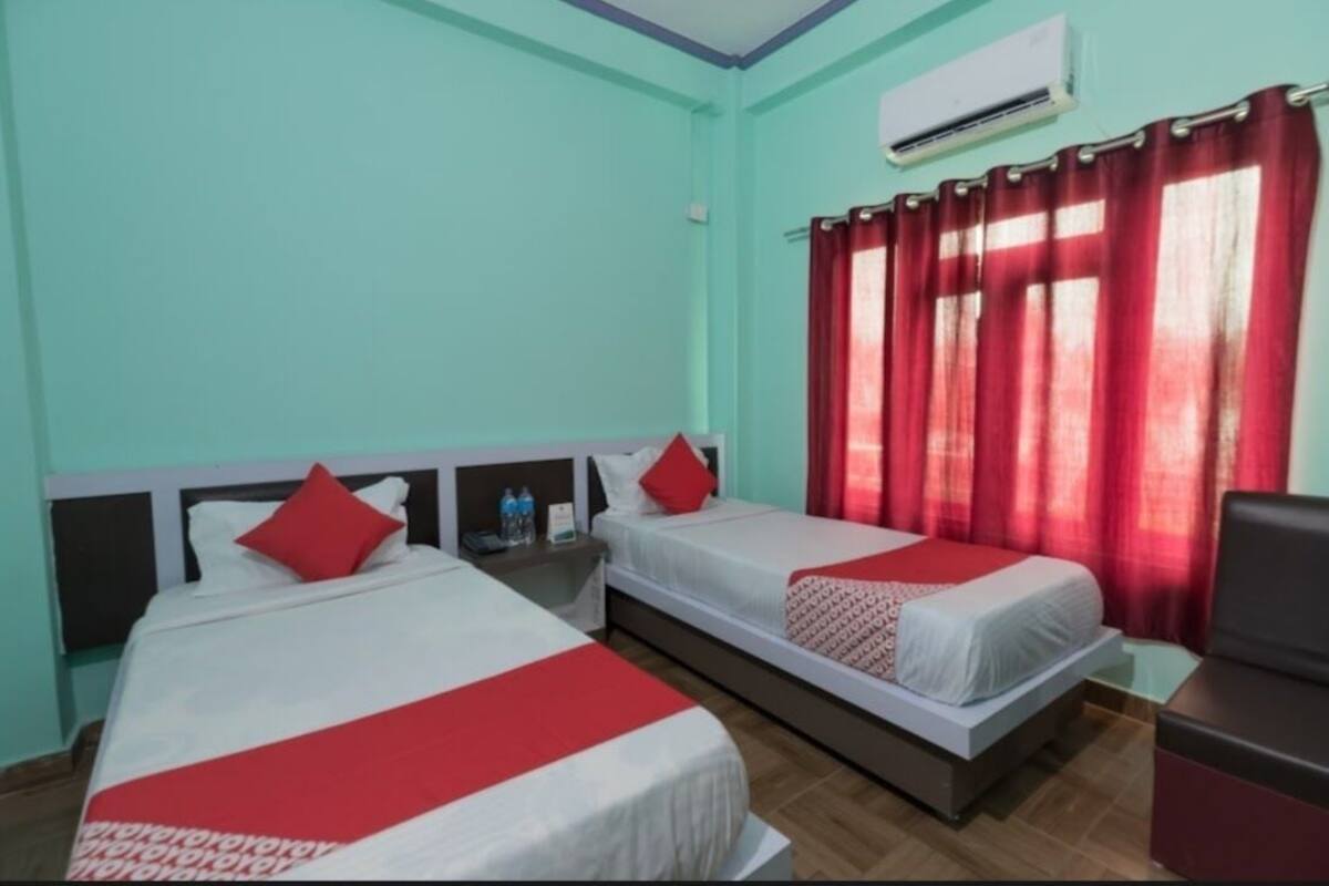 A hotel room with two beds, red curtains, an air conditioning unit, and a small table with a water bottle.