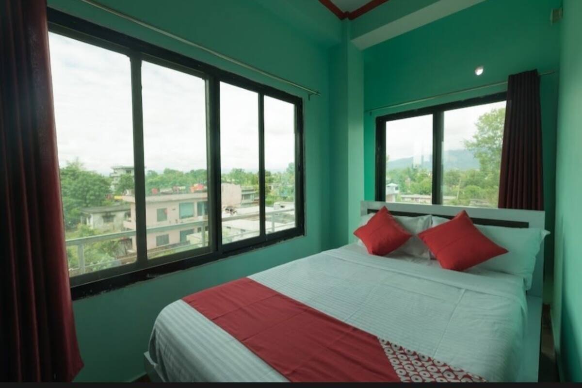 A hotel room with a large bed, two red pillows, a white headboard, and a view of buildings through large windows.