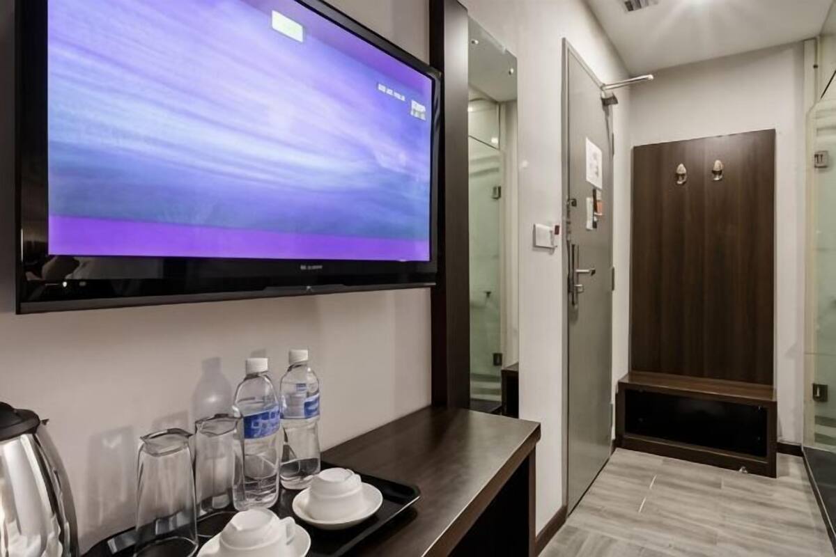 A hotel room with a flat-screen TV mounted on the wall, a wooden bench with a tray of breakfast items, and a door leading to a shower area.