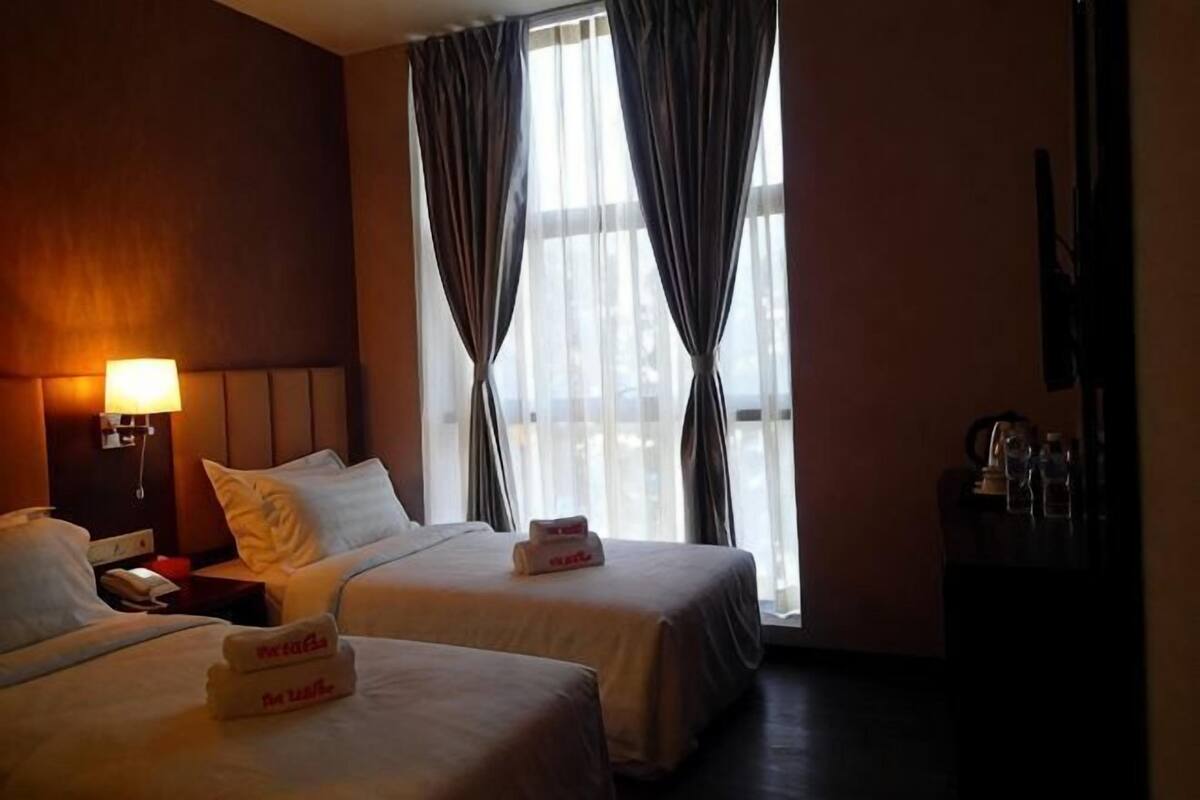 A hotel room with two beds, a bedside table with a lamp, a TV, and a window with curtains.
