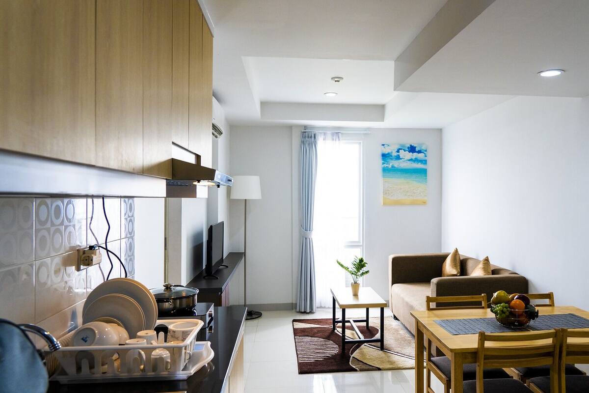 A modern living room with a sofa, a dining table with chairs, a kitchen area with a stove, and a large window with curtains.