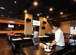 A restaurant with multiple tables and chairs, a waiter serving food, and a TV screen on the wall.