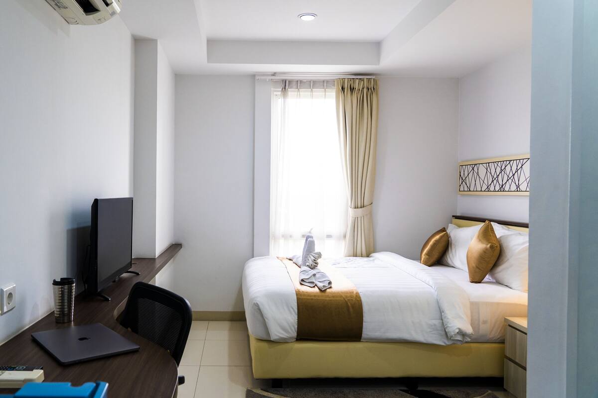 A hotel room with a bed, a desk with a laptop, a TV, and a window with curtains.