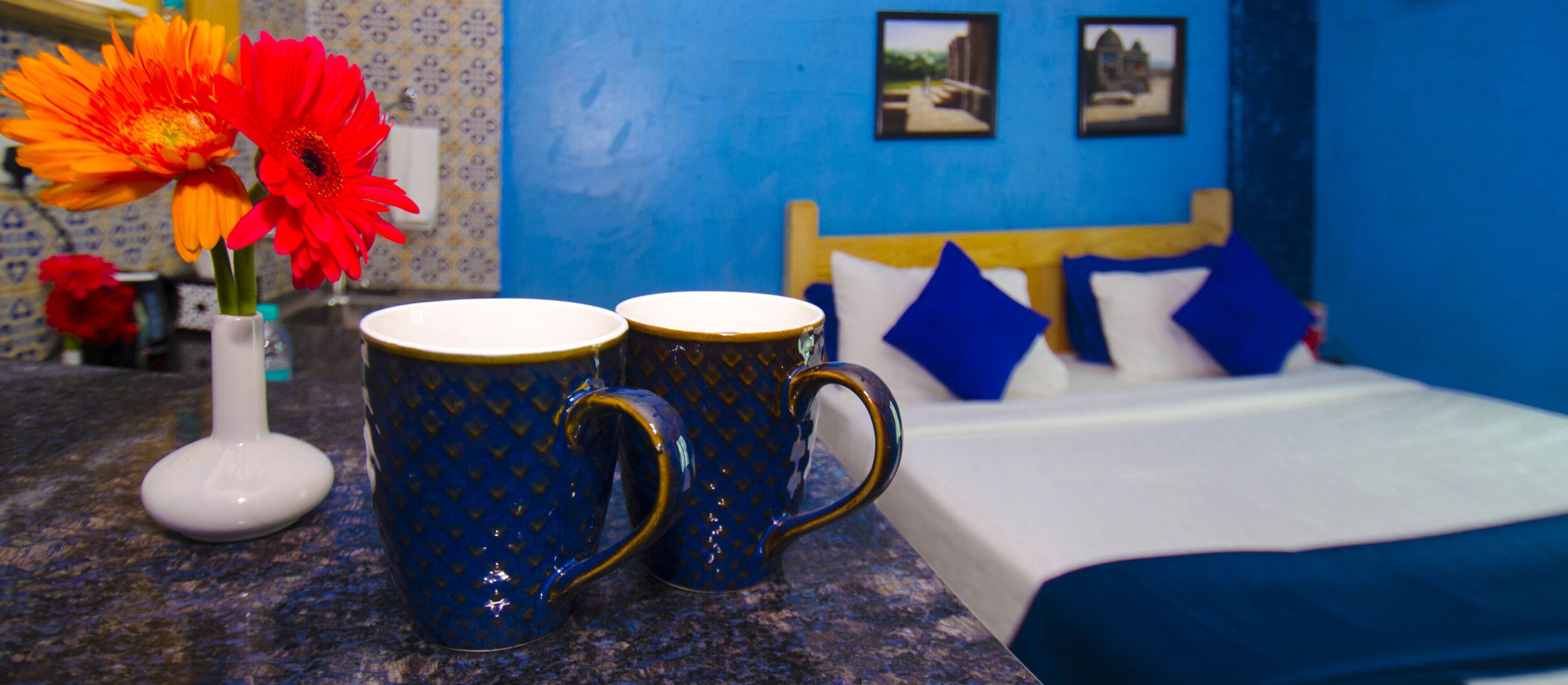 Une pièce avec un lit, deux tasses bleues posées sur un comptoir et un vase contenant des fleurs oranges.