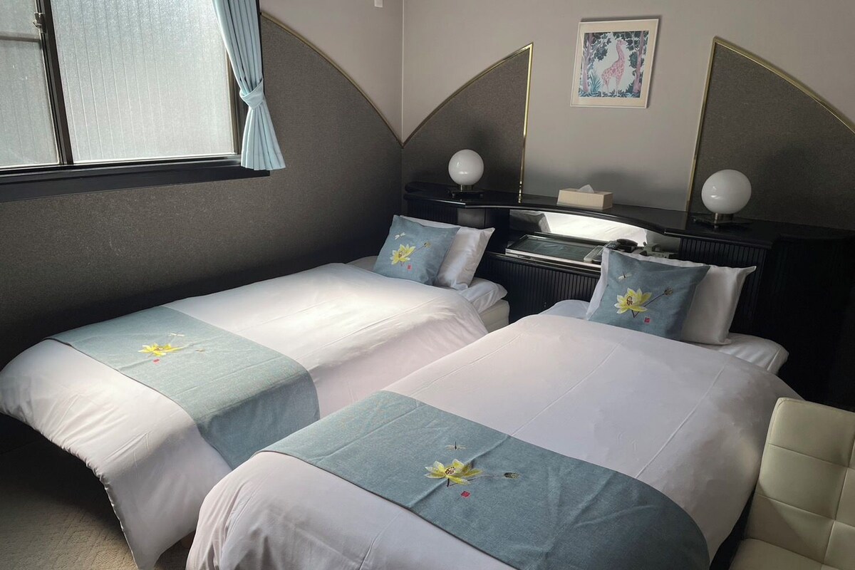 Quarto de hotel com duas camas, uma escrivaninha, uma cadeira e janela com cortinas.
