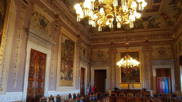 Un salón imponente con decoraciones elaboradas, una gran lámpara de araña y filas de escritorios y sillas de madera.