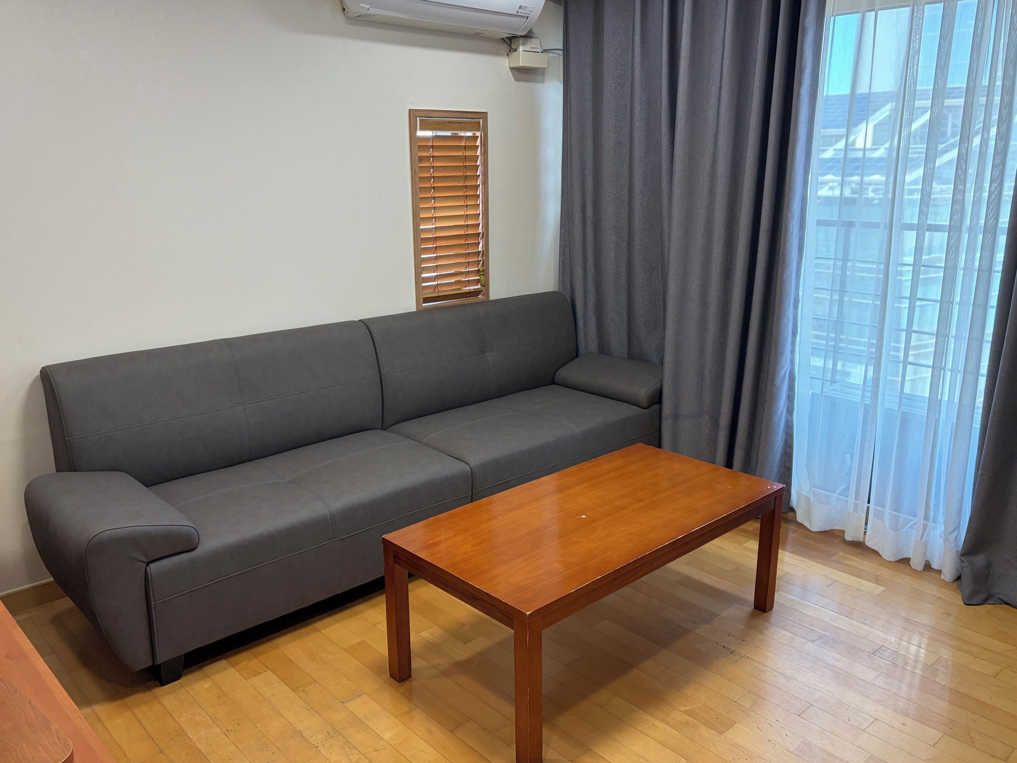 Un canapé gris, une table basse en bois et une fenêtre avec des stores et des rideaux.