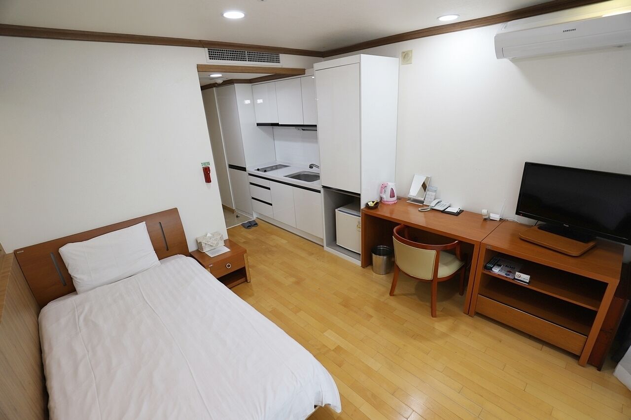 Une chambre d’hôtel compacte comprenant un lit, un bureau avec une télévision, une kitchenette et un plancher en bois.