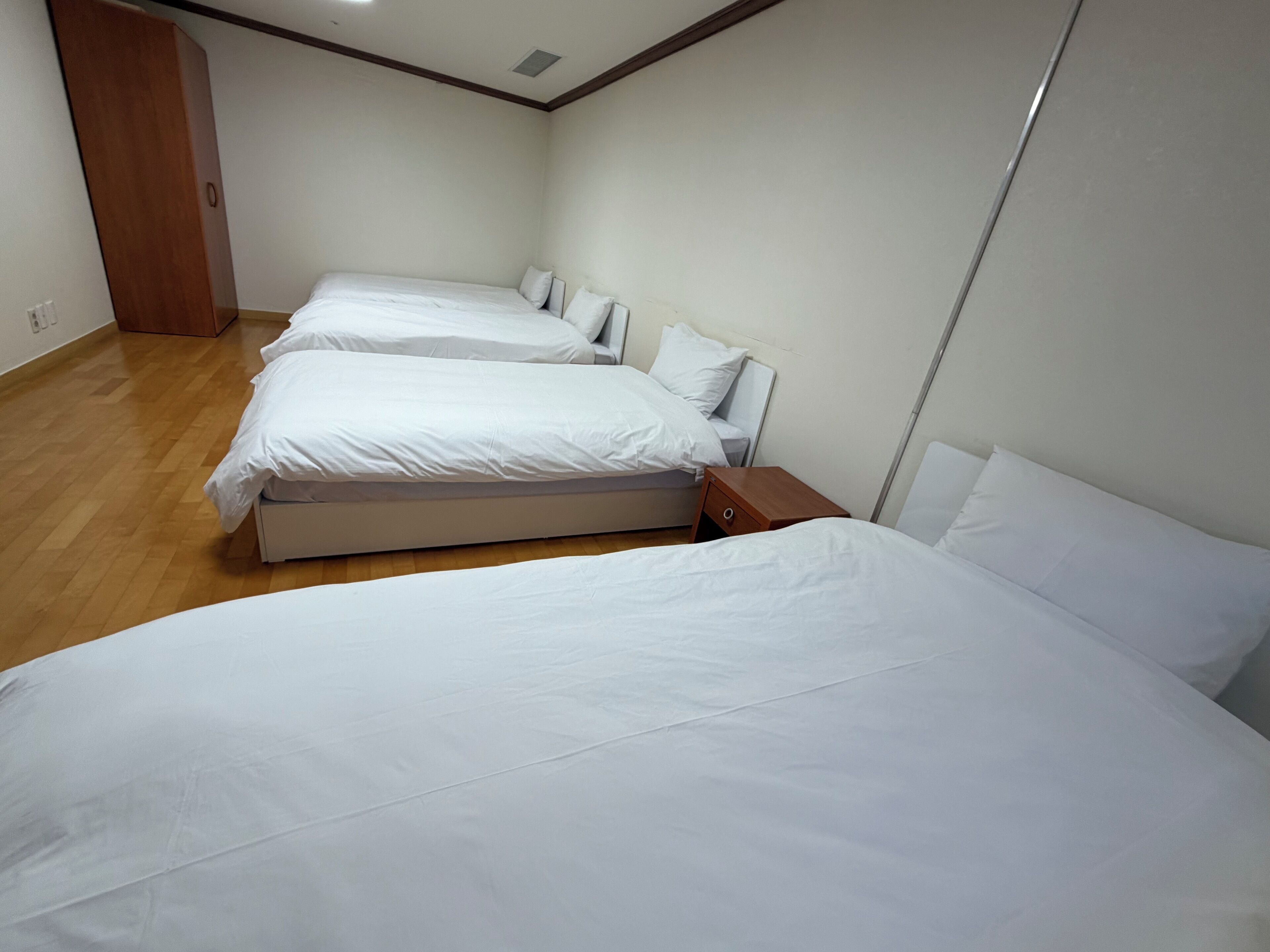 A room with three single beds aligned against a wall, a wooden wardrobe, and a wooden nightstand with a drawer.
