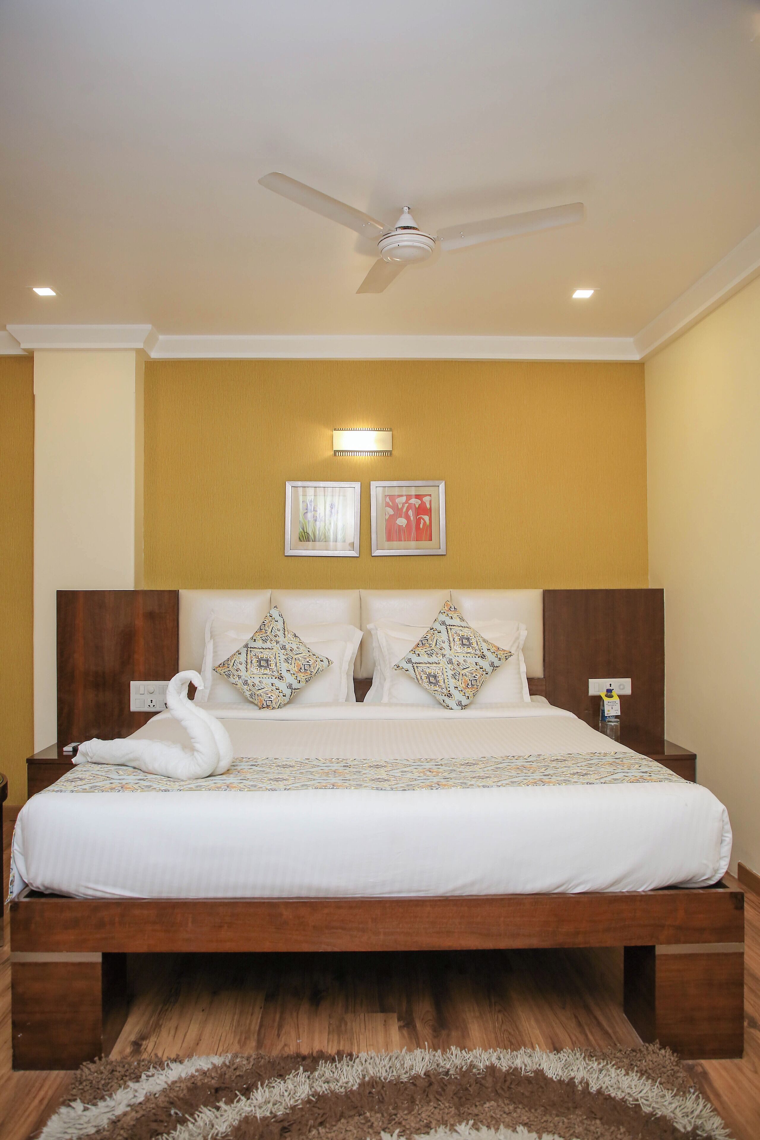 A large bed with a wooden frame, white bedding, and decorative pillows. There is a ceiling fan, wall-mounted light, and a small table with a lamp beside the bed.