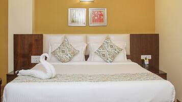 A large bed with a wooden frame, white bedding, and decorative pillows. There is a ceiling fan, wall-mounted light, and a small table with a lamp beside the bed.
