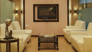 A modern living room with a flat-screen TV mounted on a patterned wall, two white sofas, a coffee table, and a chandelier. - Hotel Citrus Prime (Raipur)