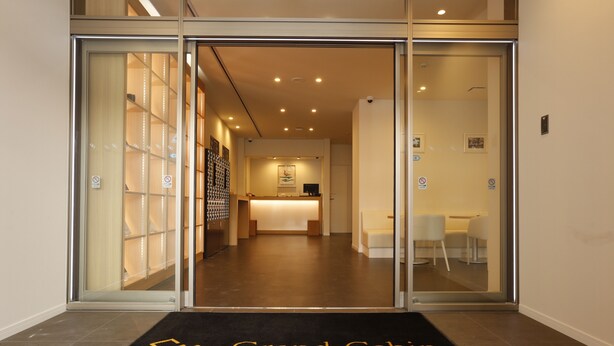Entrada de un hotel con mostrador de recepción, alfombra negra con el nombre del hotel y puerta de cristal que da a una zona de recepción.