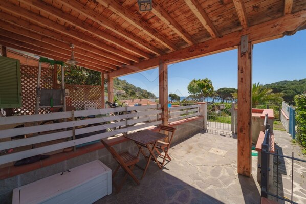 A covered outdoor seating area with a table and chairs, a white bench, and a view of the sea.