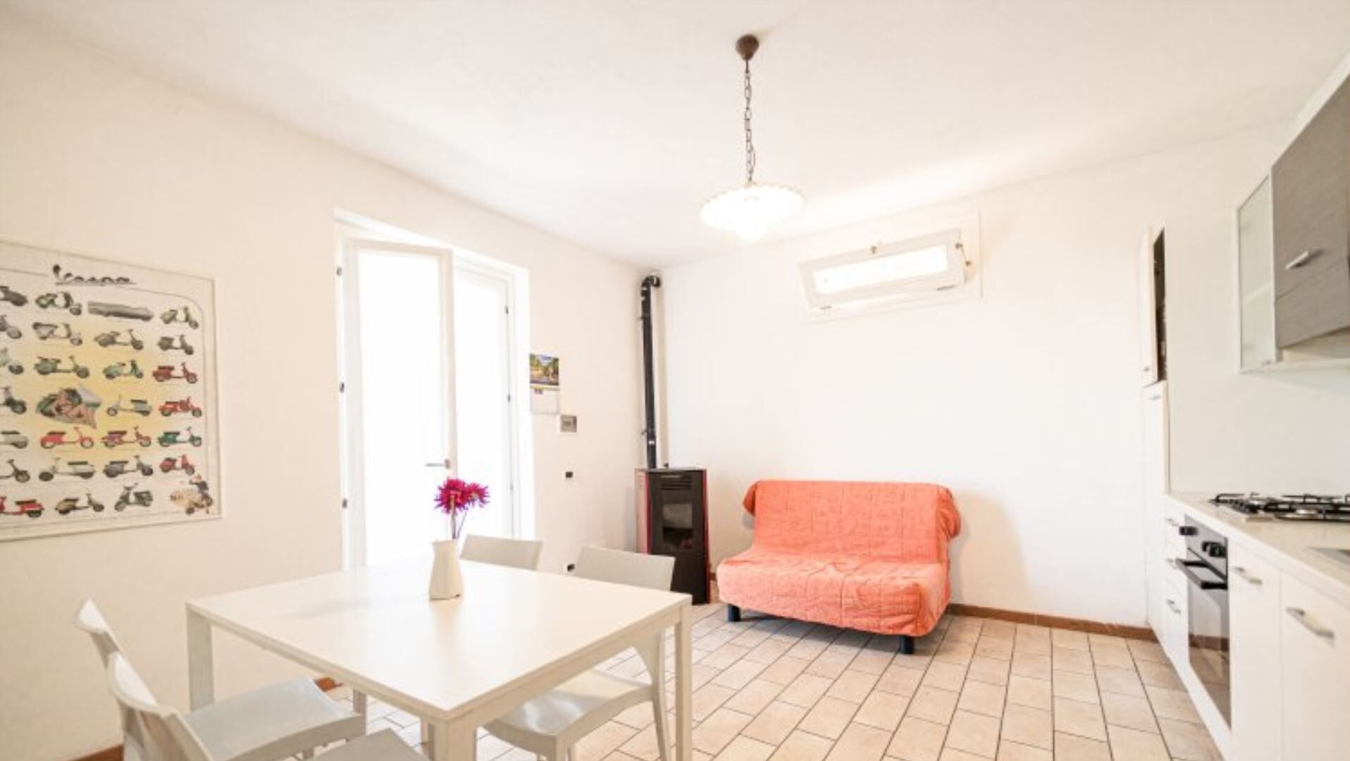 Een keuken met witte kastjes, een fornuis en een spoelbak. Er is een eettafel met stoelen en een vaas met bloemen.