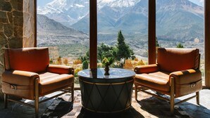 A room with two leather armchairs facing a round table with a vase of flowers. A large window offers a view of snow-capped mountains. - Shinta Mani Mustang (Jomsom)