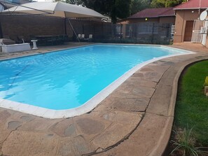 Una piscina rectangular con borde de piedra, rodeada por un muro de ladrillo y un porche cubierto con sillas.