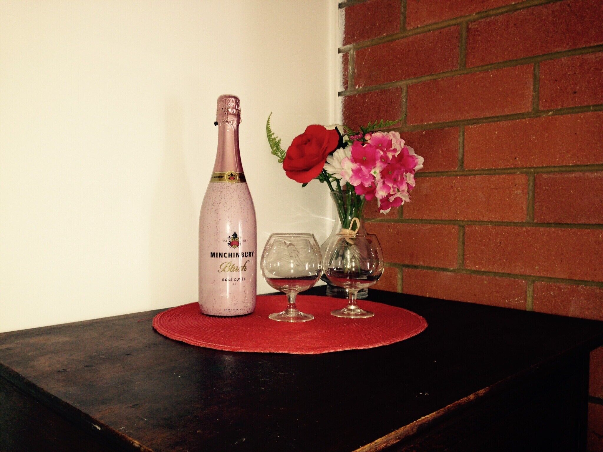A bottle of Minchinbury Blue Rosé Cuvée on a red placemat, accompanied by two wine glasses and a vase with pink flowers.