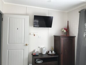 Quarto com porta branca, televisão montada, prateleira pequena com caixa de lenços, chaleira e armário com um vaso de flores.