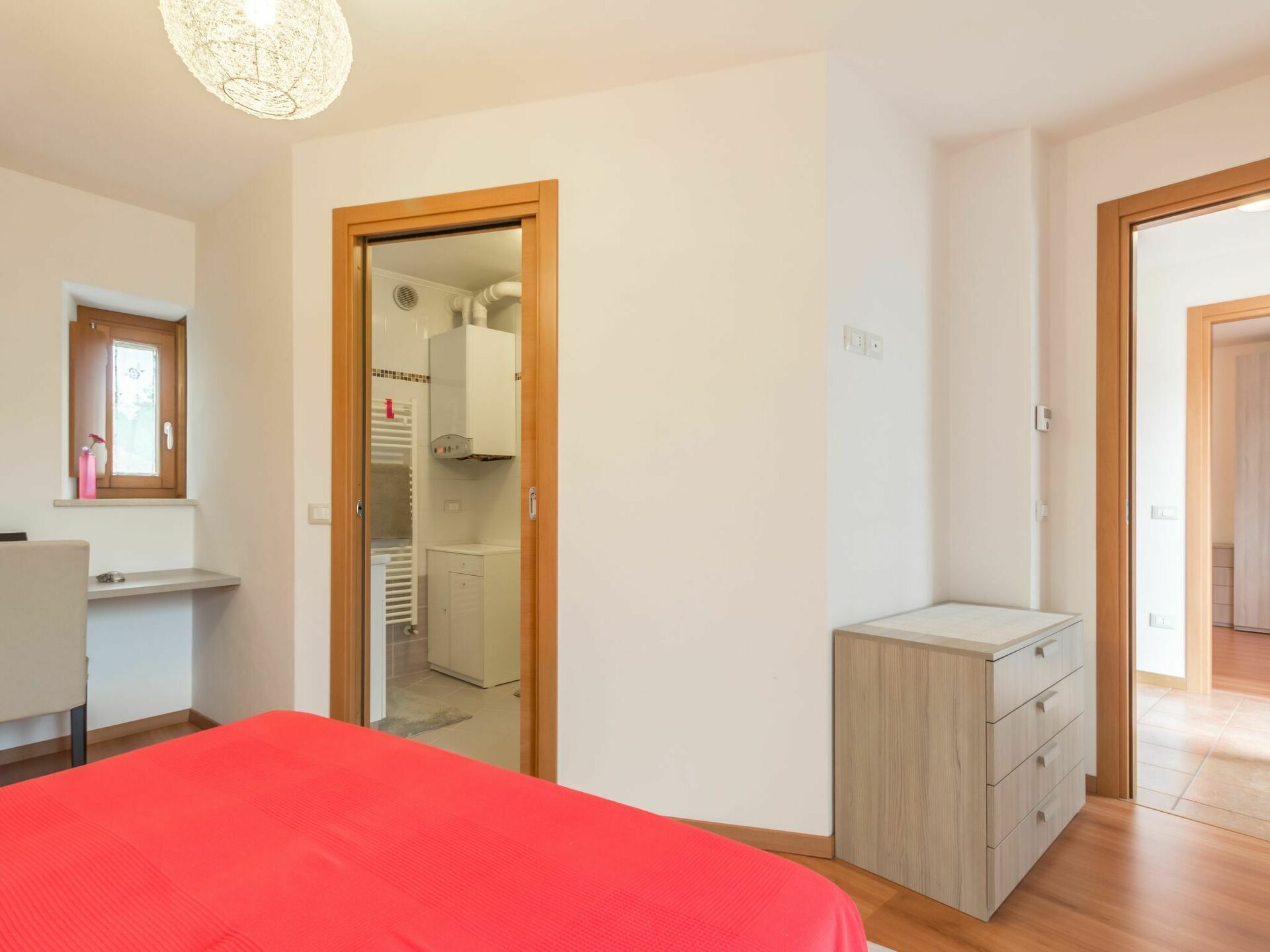 Une chambre à coucher avec un lit rouge, une commode en bois et un placard doté d’un miroir et d’une fenêtre.