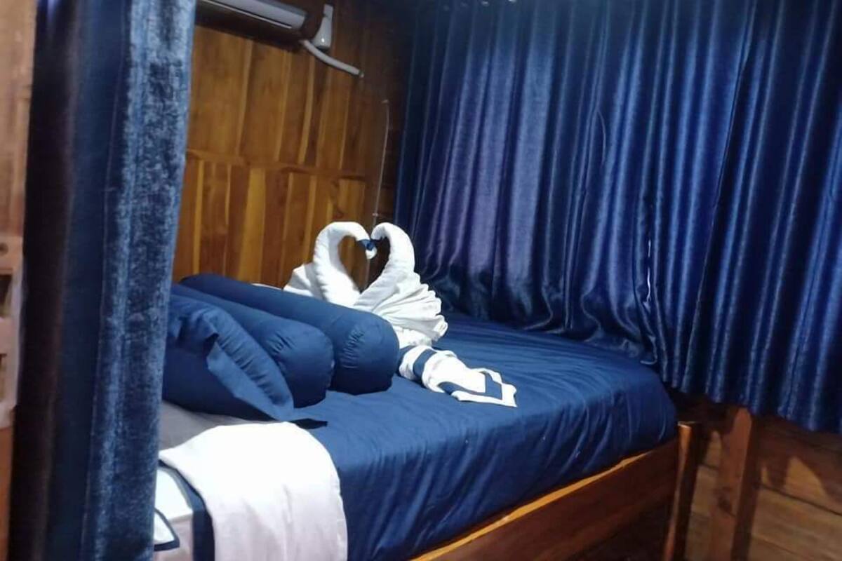 A single bed with a blue bedspread and pillows, a heart-shaped towel, and a wooden headboard.