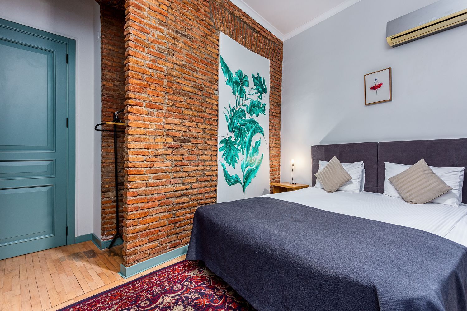 A bedroom with a brick accent wall, a bed with a grey headboard, and a red and white patterned rug.