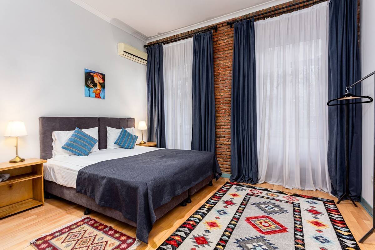 A hotel room with a bed, a nightstand, a lamp, a wall-mounted air conditioner, a brick accent wall, and a window with curtains.